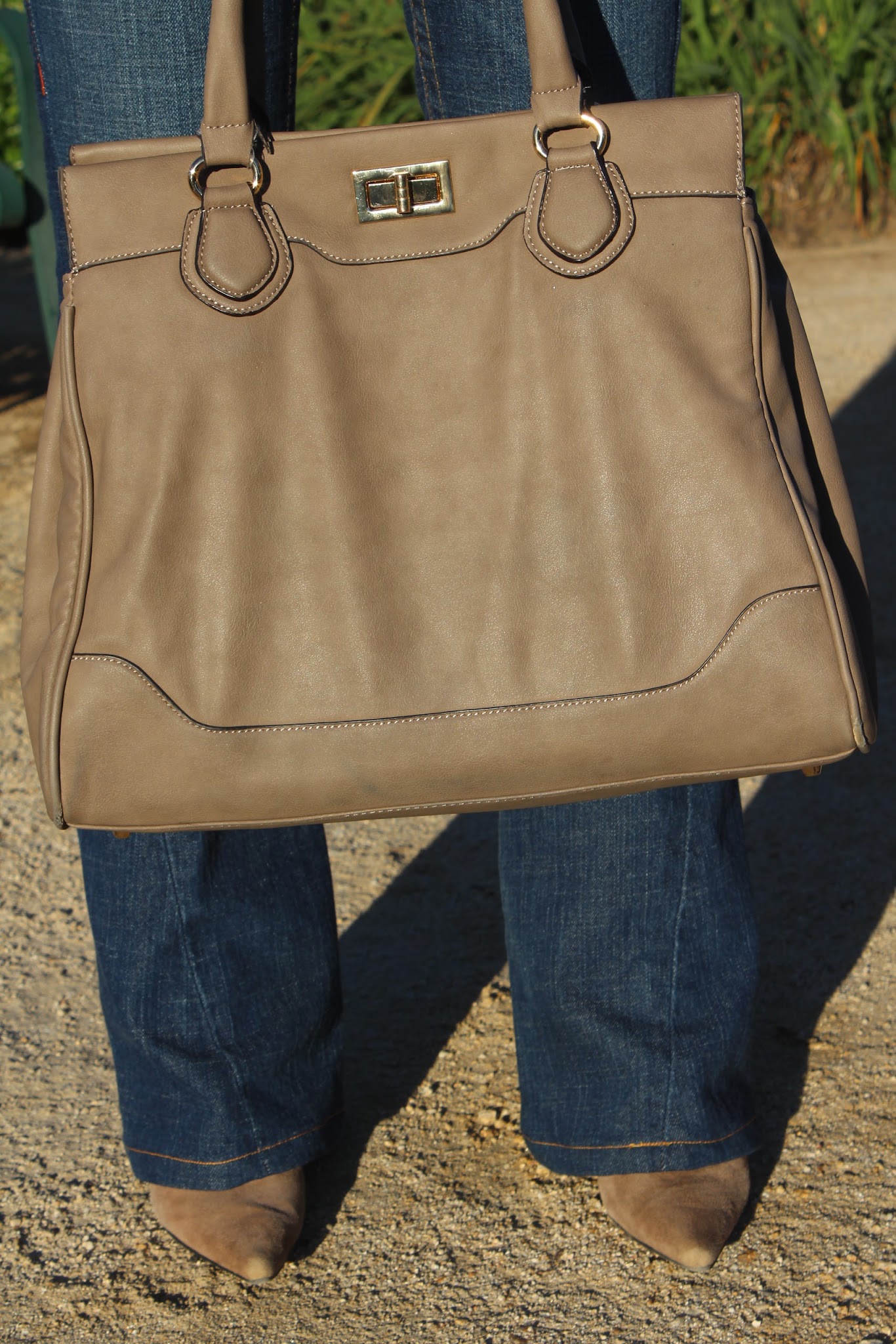 A tan leather handbag with a gold clasp, held at the side against a backdrop of soft greenery