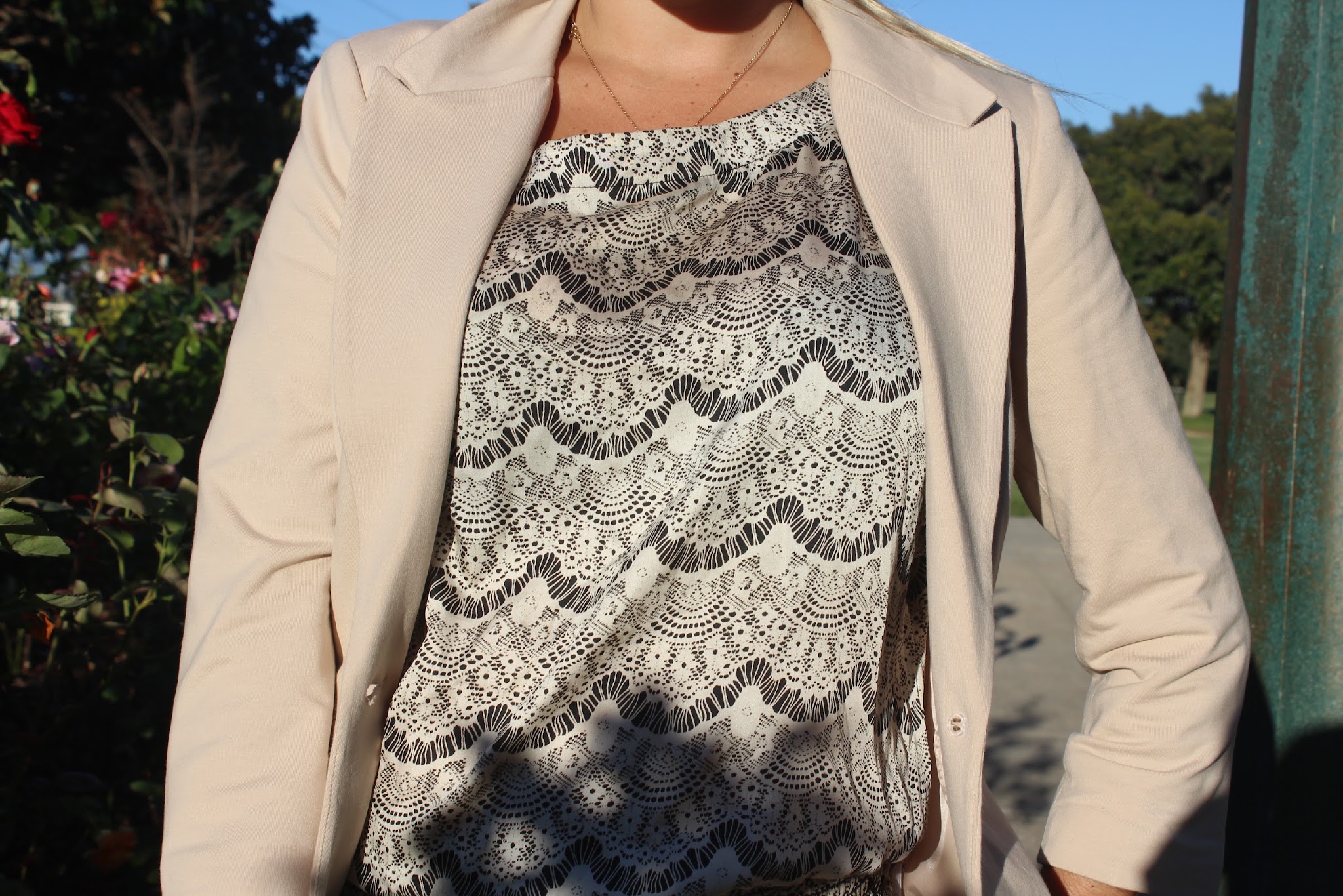A beige blazer worn over a black-and-white lace-patterned top, set against soft outdoor greenery in natural light