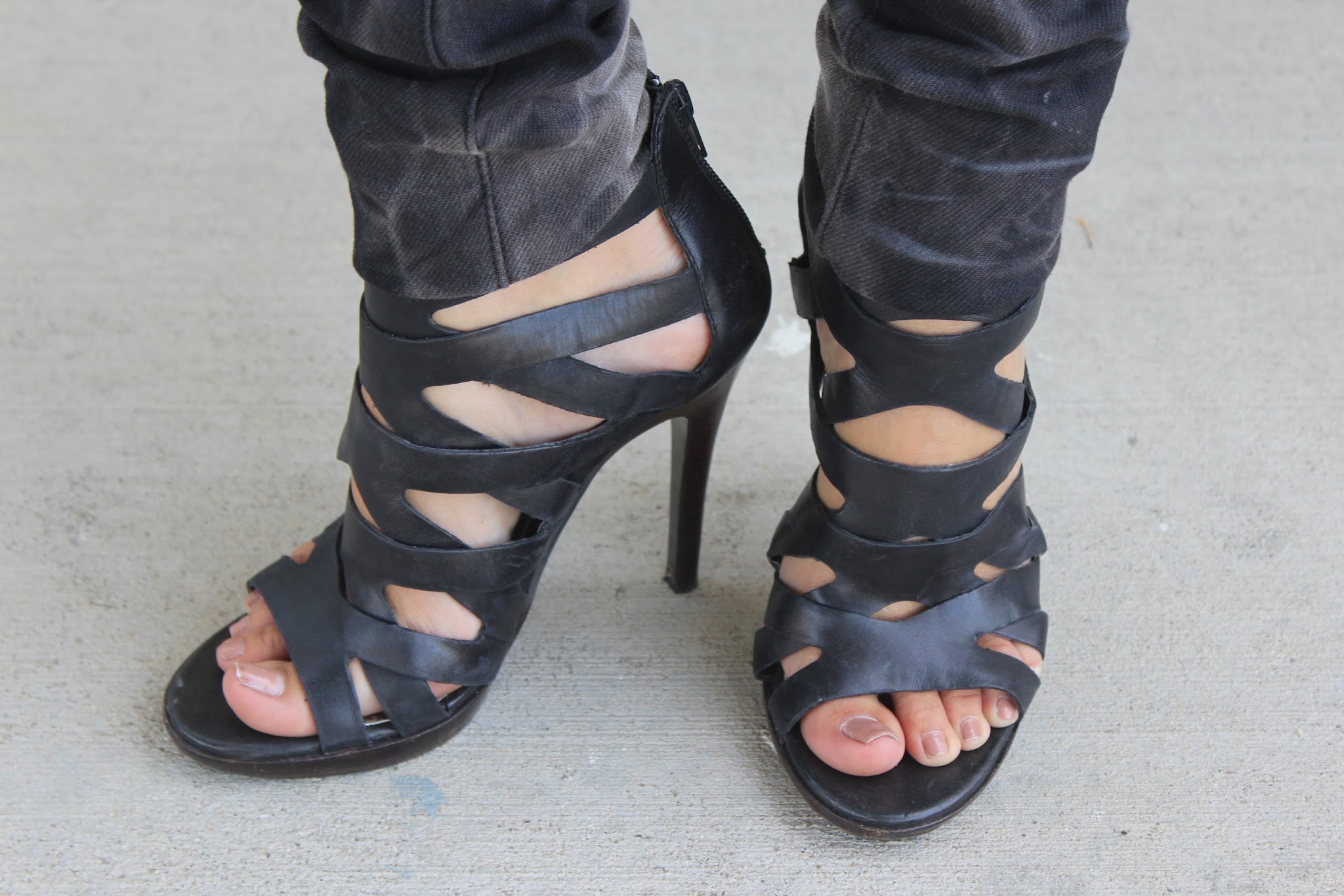 Black strappy high-heeled sandals with cuffed dark jeans, standing on a concrete surface
